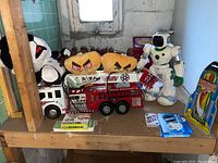 Photo showing a large plastic firetruck toy, multiple Angry Birds stuffed toys and panda plush, white robot toy, and several card games on a wooden shelf in a basement.