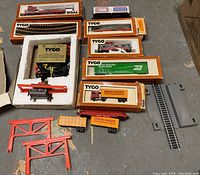 Overview of boxed and loose TYCO HO scale train cars, track pieces, power supply, and accessories arranged on concrete floor.