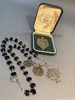 Full lot view showing black onyx bead necklace with attached religious charms laid out beside the Sacred Heart heart pendant in green Harrod's jewelry box.
