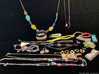 Overview of multiple boho chic style necklaces and bracelets arranged on dark surface, showing varied colors and styles including yellow beads, multicolor beads, metal accents, and flower accessory.