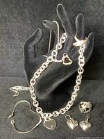 Seven pieces of sterling silver jewelry arranged on a black hand display including chains, rings, and earrings showcasing heart motifs and different silver chain styles