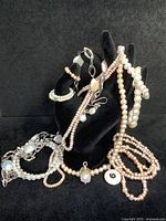 Overall view of various pearl necklaces, bracelets, rings, and earrings displayed on a black stand and surface highlighting different pink and white pearls with metal clasps and pendants.