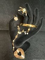 Six jewelry items displayed on black hand stand: ring, earrings, froggy brooch, heart brooch, gold bangle, and filigree pearl bracelet.