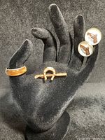 Photo of black mannequin hand displaying all three items: gold horseshoe and crop pin on middle finger, tiger's eye ring on ring finger, and horse head cufflinks attached to index finger.