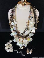 Photo of 6 pieces of pearl jewelry displayed on mannequin including several multi-strand necklaces with various colored pearls and large mother of pearl disc decorations, a brooch shaped like flower buds on stems, a pearl bracelet, and drop earrings.