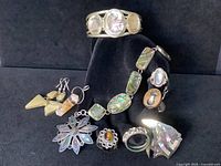 All jewelry pieces displayed on black background, including rings, a flower ring with tiger's eye, abalone inlay bracelets, brooches, pendant, and earrings.