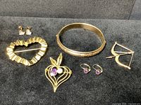 Full view of six jewelry pieces laid out on black fabric including heart post earrings, gold and amethyst drop earrings, bangle, and three brooches including a bow and arrow design