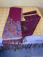 Two folded vibrant wool throws, one in deep purple and another in a darker multi-color paisley design with fringed edges on a beige surface.