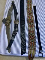 All five belts laid out side by side on a striped background, showing varying widths and buckle styles.