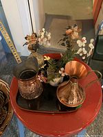 Top perspective of five items: two Turkish copper jars (one pitcher-style with handle, one cylindrical with engraving) and three small glass jars with dainty glass flowers that have white, pink, and light orange petals; all on a black tray on a red table.
