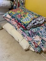 Stack showing folded duvet with floral and geometric colorful pattern on top, solid cream bedskirt beneath, and quilt/comforter with a similar floral pattern below.