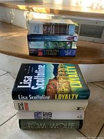 Stack of hardcover books on wooden steps featuring various titles by Tom Wolfe, Lisa Scottoline, James Clavell, Rosie Thomas, Edwina Currie, Lana Kortchik, and Frederick Forsyth.