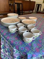 Full display of the dishes including stacked dinner plates, side plates, bread plates, teacups arranged in front on a floral tablecloth.
