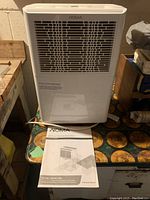 White Noma dehumidifier with front view showing air vent grill and placed on table with manual in front.