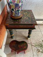 Side view of the painted wooden side table with turned green legs accented with yellow and a small heart-shaped painted stool on tile floor.