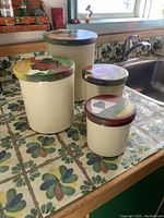 Four cream-colored canisters on tiled kitchen counter with colorful hand-painted wooden lids, shown from a side angle.