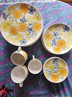 Photo showing 2 stacks of plates and 4 teacups with yellow and blue floral print on white porcelain.