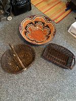 Photo showing the three darker wicker baskets arranged on a carpeted floor: a large round basket with colorful geometric pattern and two smaller dark wicker baskets, one oval handled and one rectangular with handles.