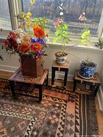 Three small wooden stools arranged near a window; the largest holds a copper tin with fabric flowers, two others hold potted plants.