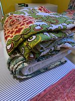 Folded view of two comforters stacked, showing bold floral and swirl print in bright green, orange, and white on top and a muted patterned comforter beneath.