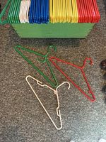 Three plastic hangers in green, red, and cream outside the crate on carpeted floor to show shape and size alongside the crate with hangers inside.