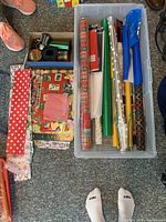 Top view of plastic bin packed with multiple rolls of varied wrapping paper and tissue paper, accompanied by a box with ribbons and small accessories beside it.