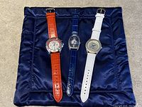 Front view of three Akribos watches placed on a dark blue satin cloth, showing face designs and leather straps in orange, blue, and white colors.