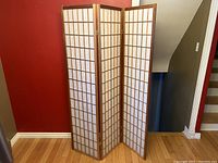 Front view of the three-panel wood and paper room divider set against a red and gray wall with hardwood flooring, showing the unbent position.