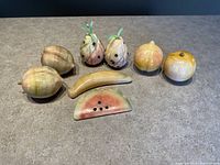 Full view of all eight alabaster stone fruit figurines including 2 pineapples, 2 pears, 2 peaches, 1 banana, and 1 watermelon slice on grey surface.