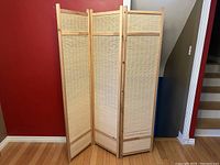 Full front view of the closed 3-panel room divider showing woven bamboo reed panels within light wood frames against a red wall and hardwood floor.