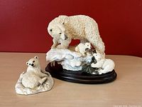 Photo of two polar bear figurines against a red background. The larger figurine features an adult bear with two cubs on a snowy base with wooden stand; the smaller figurine is a sitting cub on its own base.