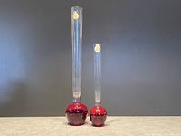 Two vintage Holmegaard red bubble bud vases, one taller and one shorter, with clear elongated necks and red bubble bases, standing together on a gray surface against a dark background.