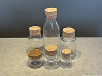 Photo of six transparent glass jars with cork lids arranged in two rows by size on a countertop against a dark background.