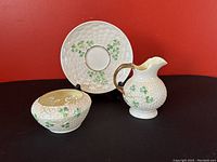 Three-piece set showing Irish Belleek cream pitcher with gold-toned handle, sugar bowl with crackled glaze interior, and saucer plate decorated with shamrocks, all with textured basket-weave pattern.