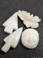 Four carved opal pieces in white tone with subtle iridescence. Two arrowhead shapes, one floral carving, and one face sculpture.