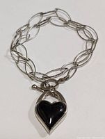 Close-up photo of silver necklace with elongated oval links and black onyx heart pendant attached near toggle clasp.