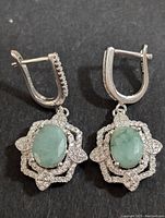 Front view of the pair of silver earrings with green oval emerald stones and surrounding cubic zirconia in floral design.