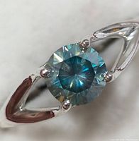 Close-up image showing the 1.35ct round-cut blue moissanite gemstone set in a silver band with prongs.