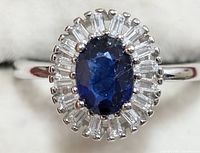 Top view of silver ring featuring oval blue sapphire center with surrounding clear baguette-cut cubic zirconia stones.