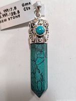 Front angled view of silver turquoise pendant showing natural turquoise hexagonal prism and ornate silver top with round cabochon gemstone.