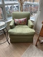 Green upholstered armchair positioned by a window, with a floral decorative cushion on the seat.