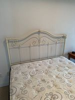 White iron queen-size bedboard against a wall with mattress beneath, showing ornate scrollwork design and finials at top corners.