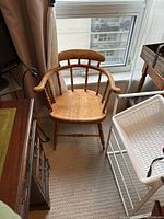 Pine wood armchair positioned near a window, surrounded by various furniture and household items. The chair displays a curved backrest with spindle supports and curved armrests. Light natural wood finish.