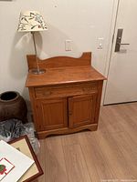 Photo of a pine wood side table with a drawer and cabinet doors placed against the wall, with a lamp on top.