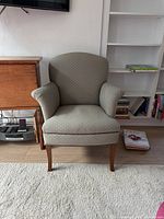 Front view of a green upholstered armchair with wooden legs, pictured in a room with bookshelf, TV, and wooden side table.