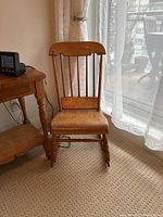 Front view of pine wooden rocking chair with spindled backrest and flat wooden seat, placed on carpet near a wooden table and window with curtain.