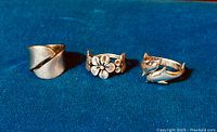 Three sterling silver rings on blue fabric background, showing front designs including the wide band, floral, and fox-shaped rings.