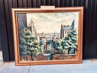 Full view of oil painting in wooden frame with mounted light, showing cityscape with trees and buildings.