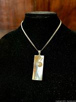Full view of the pendant necklace displayed on black stand showing rectangular mother-of-pearl pendant with Mabe pearl and silver chain.