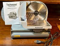 Full view of the Techwood electric food slicer showing blade, handle, base, and power cord with instruction manual behind.